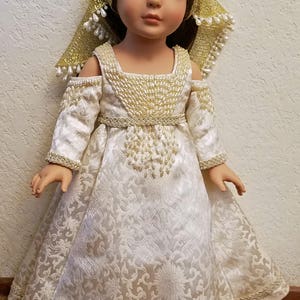 Puede incluir: Una muñeca blanca con un vestido blanco con ribetes dorados y un pañuelo de cabeza dorado con cuentas blancas. El vestido tiene un estampado floral y la muñeca tiene ojos azules.