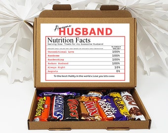 May include: A brown cardboard box with a white label that says "Awesome Husband Nutrition Facts" and lists various qualities with a percentage next to each. The box is open and contains a variety of chocolate bars.