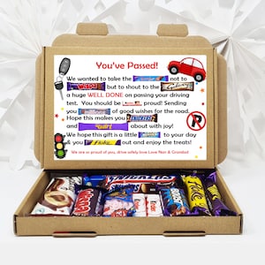 May include: A brown cardboard gift box containing assorted chocolate bars. The box lid features a message, "You've Passed!", with car and key graphics. The box is open, displaying a selection of chocolate treats inside, perfect for a celebration.