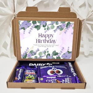 May include: A brown cardboard box with a floral design and the text "Happy Birthday" on the lid. The box is open and contains a variety of chocolate bars, including Dairy Milk, Freddo, and Cadbury.