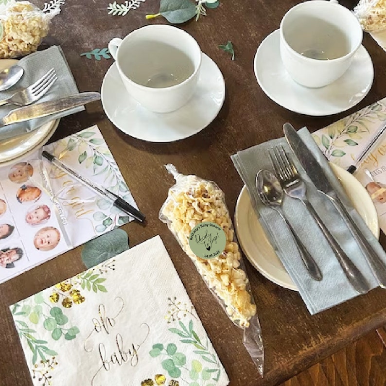 May include: A table setting for a baby shower with a green and gold theme. There are two teacups and saucers, a napkin with the words "oh baby" printed on it, a bag of popcorn with a sticker that says "Daddy's Baby Boy!" and a pen on a sheet of paper with a list of names.