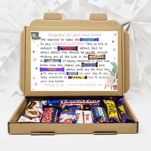 May include: A brown cardboard box with a pink and white card inside. The card has a message that says "Congrats on your new home!" and is decorated with images of chocolate bars. The box is open and filled with various chocolate bars.