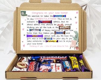 May include: A brown cardboard box with a pink and white card inside. The card has a message that says "Congrats on your new home!" and is decorated with images of chocolate bars. The box is open and filled with various chocolate bars.