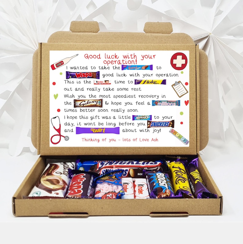 Puede incluir: Una caja de cart&oacute;n marr&oacute;n con la tapa abierta, que revela una variedad de barras de chocolate. La caja tiene un mensaje escrito a mano en el interior que dice "&iexcl;Mucha suerte con tu operaci&oacute;n!" e incluye una lista de nombres de barras de chocolate como "Wispa", "Galaxy", "Twirl", "Boost" y "Snickers".