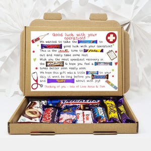 Puede incluir: Una caja de cart&oacute;n marr&oacute;n con la tapa abierta, que revela una variedad de barras de chocolate. La caja tiene un mensaje escrito a mano que dice "&iexcl;Mucha suerte con tu operaci&oacute;n!" e incluye una lista de nombres de barras de chocolate, como "Wispa", "Galaxy", "Twirl" y "Boost".