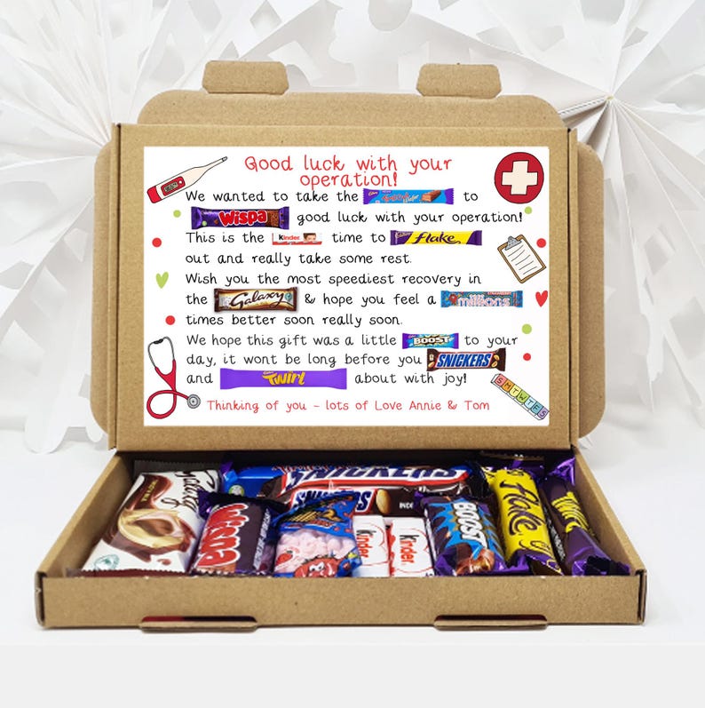 Puede incluir: Una caja de cart&oacute;n marr&oacute;n con la tapa abierta, que revela una variedad de barras de chocolate. La caja tiene un mensaje escrito a mano que dice "&iexcl;Mucha suerte con tu operaci&oacute;n!" e incluye una lista de nombres de barras de chocolate, como "Wispa", "Galaxy", "Twirl" y "Boost".