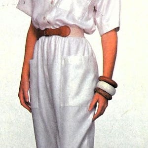 May include: A white jumpsuit with a button-up front and short sleeves. The jumpsuit has a brown belt and a pocket on each leg. The model is wearing the jumpsuit with a pair of black shoes.