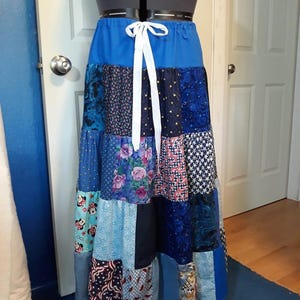 Op de afbeelding: Een lange patchwork rok in blauwtinten, met verschillende patronen en texturen. De rok heeft een effen blauwe tailleband met een wit trekkoord. Het patchwork bevat bloemen-, ster- en geometrische ontwerpen, waardoor een uniek en kleurrijk kledingstuk ontstaat.