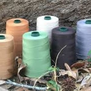 May include: Seven spools of thread in various colors, including brown, beige, green, black, and gray. The spools are arranged in a row on a bed of leaves.