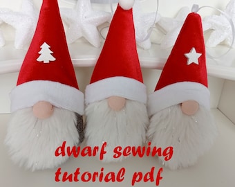 Può includere: Tre decorazioni natalizie a forma di gnomi con cappelli di velluto rosso, barbe bianche e nasi beige. Ogni gnomo ha un bordo bianco sul cappello e un piccolo abbellimento bianco: un albero, un pompon e una stella. Il testo "dwarf sewing tutorial pdf" è scritto in rosso.