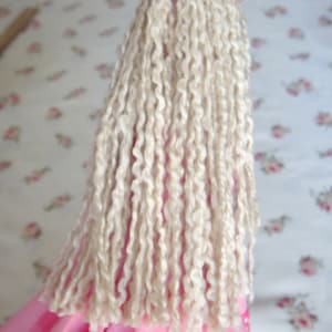 May include: A handmade doll with long, white yarn hair and a pink dress. The doll is wearing a pink flower on its head.