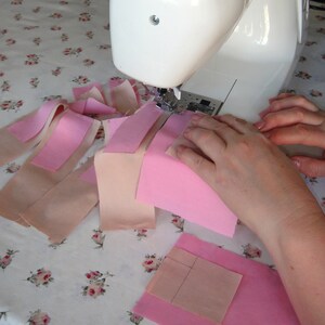 May include: A white Singer sewing machine is being used to sew together pink and beige fabric scraps. The fabric scraps are arranged in a pattern on a white tablecloth with a floral design.