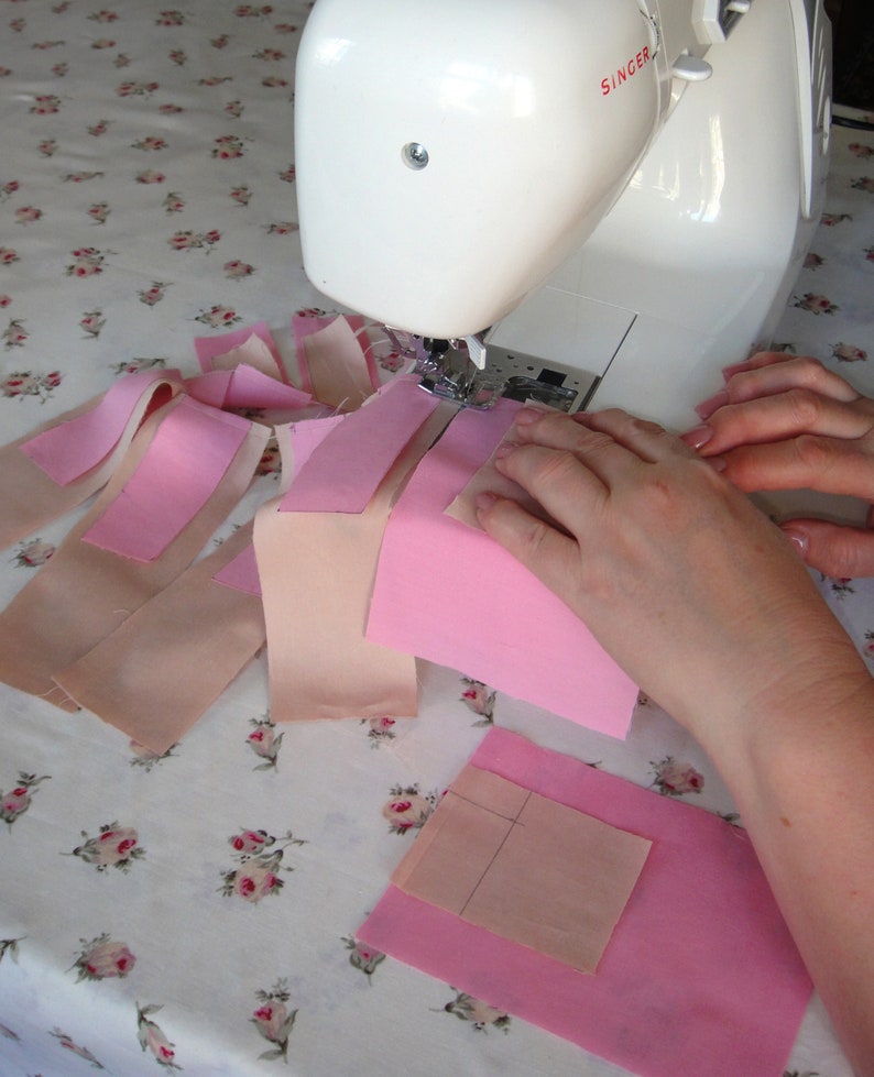 May include: A white Singer sewing machine is being used to sew together pink and beige fabric scraps. The fabric scraps are arranged in a pattern on a white tablecloth with a floral design.