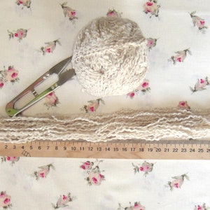 May include: A ball of beige yarn, a ruler marked with numbers from 0 to 30, and a pair of scissors on a white fabric background with a floral pattern.