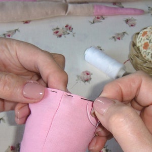 May include: Close-up of a person's hands sewing a pink fabric with a needle and thread. The fabric is folded in half and the person is stitching along the edge. A spool of white thread and a pincushion are visible in the background.