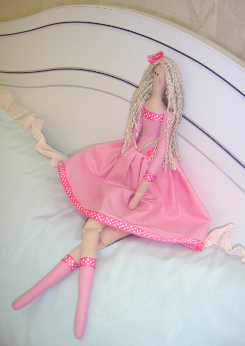 May include: A handmade doll with long blonde hair, wearing a pink dress with white polka dots and a pink ribbon. The doll is sitting on a light blue bedspread.