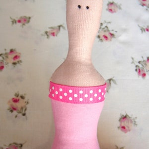 May include: A pink fabric doll with a white polka dot ribbon around its waist. The doll has a pink dress and a black hair bow.