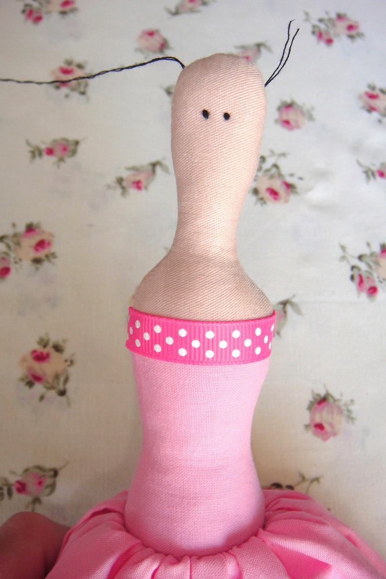 May include: A pink fabric doll with a white polka dot ribbon around its waist. The doll has a pink dress and a black hair bow.