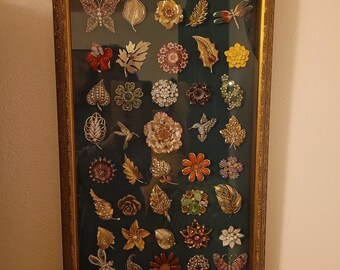 May include: A framed display of 36 vintage brooches with a variety of designs, including butterflies, flowers, and geometric shapes. The brooches are arranged in a grid pattern and are set against a dark background.