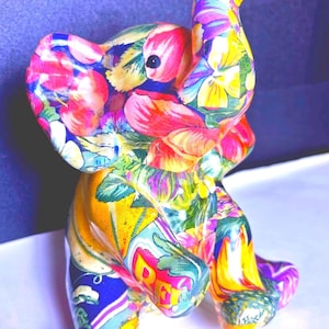 May include: A colorful ceramic elephant figurine. The elephant is covered in a vibrant floral pattern with red, pink, yellow, and blue hues. The trunk is raised, and the ears are large. The figurine is standing on a white surface.