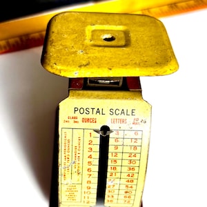May include: Vintage yellow postal scale with a rectangular base and a weighing platform. The scale has a black indicator and a chart for ounces and letters. The text "POSTAL SCALE" is visible.