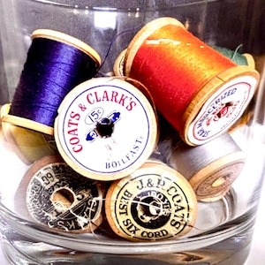 May include: A clear glass jar filled with vintage spools of thread. The spools are various colors, including purple, orange, and gray, with labels from Coats & Clark's and J&P Coats. The thread spools are a mix of sizes and styles.