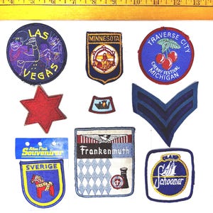 May include: A collection of embroidered patches with various designs. Included are patches with the text "LAS VEGAS", "MINNESOTA", "TRAVERSE CITY", "Frankenmuth", and "Schooner". Other patches feature a red star, a shield with a horse, and a blue chevron.