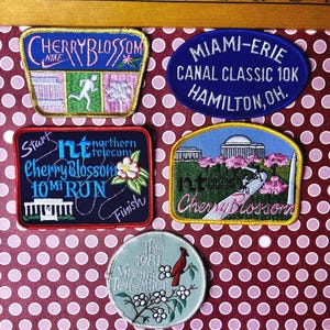 May include: A collection of embroidered patches with various designs and text. The patches include the words "Cherry Blossom," "Miami-Erie Canal Classic 10K," and "Virginia Ten Miler." Colors include blue, pink, green, and yellow.