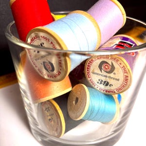May include: A clear glass filled with colorful spools of thread. The spools are various colors, including red, light blue, orange, purple, and teal. The spools have wooden cores and are labeled with text including "American thread".