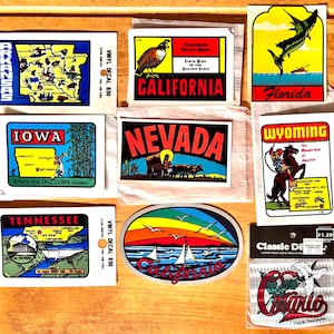 May include: A collection of vintage vinyl decals featuring state-themed designs. The decals showcase illustrations of Arkansas, California, Florida, Iowa, Nevada, Tennessee, Wyoming, and Ontario. Each decal has a unique design, including maps, state symbols, and scenic views.