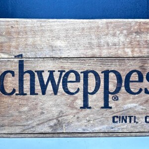 May include: A weathered wooden crate with the word "Schweppes" in large, dark letters. The text "CINTI. OHIO" is printed below. The crate has a rustic, aged appearance, with visible wood grain and a faded color.