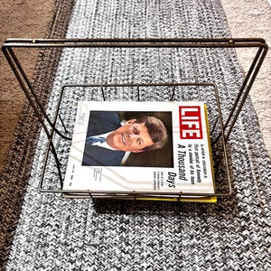 May include: A vintage metal magazine rack holding a copy of LIFE magazine. The magazine cover features a portrait of John F. Kennedy. The rack is made of thin, gold-toned metal.