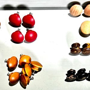 May include: A collection of various cabinet knobs in different styles and colors. The knobs include wooden, red, and black designs, with some featuring metal hardware for attachment. The knobs are arranged on a white surface.