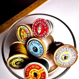 May include: A collection of vintage wooden spools of thread in a clear glass bowl. The spools have colorful labels with text like "Barbours Linen Thread" and "J&P Coats Heavy Duty". A wooden ruler is in the background.
