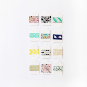 May include: A collection of white paper tags with decorative washi tape. The tapes feature various patterns, including floral, geometric, and chevron designs. The color palette includes pink, blue, yellow, and gold, creating a visually appealing assortment for crafting and gift wrapping.