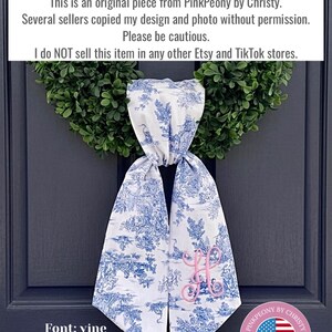 K&ouml;nnte beinhalten: Ein hellblaues Toile-de-Jouy-T&uuml;rschal aus Leinen mit einem rosa Monogramm, das an einer dunkelblauen T&uuml;r h&auml;ngt. Der Schal hat ein blau-wei&szlig;es Pastoraldesign. Der Text "PinkPeony by Christy" steht oben.