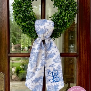 K&ouml;nnte beinhalten: Ein wei&szlig;-blau gemusterter Schal, ca. 137 cm lang, mit einem blau gestickten Initial. Der Schal wird an einer Glast&uuml;r mit einem gr&uuml;nen Kranz pr&auml;sentiert. Der Text "PinkPeony by Christy" steht oben.