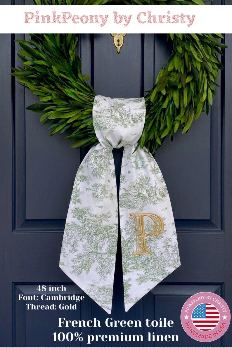 K&ouml;nnte beinhalten: Eine wei&szlig;e und franz&ouml;sisch-gr&uuml;ne Toile-de-Jouy-T&uuml;rsch&auml;rpe mit einem goldgestickten Buchstaben "P". Die Sch&auml;rpe ist 122 cm lang und aus 100 % Premium-Leinen gefertigt. Die Sch&auml;rpe ist um einen gr&uuml;nen Kranz an einer marineblauen T&uuml;r gebunden.