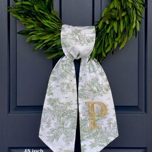 以下が含まれることがあります： 金色の刺繍されたイニシャル「P」が付いた、白とフレンチグリーンのトワルドジュイのドアサッシュ。 サッシュの長さは122cmで、100％プレミアムリネン製です。 サッシュは、濃い青色のドアの緑色の花輪に結ばれています。