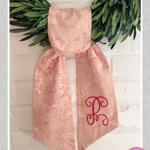 K&ouml;nnte beinhalten: Ein rosa Toile-Schal mit einem rot gestickten Initialen. Der Schal ist aus rosa Stoff mit franz&ouml;sischem Cameo-Design gefertigt. Der Text "PinkPeony by Christy" steht oben.