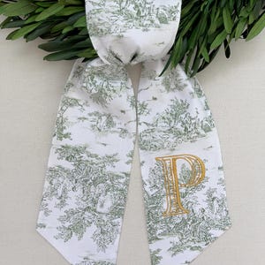 K&ouml;nnte beinhalten: Wei&szlig;er Schal mit gr&uuml;nem Toile-de-Jouy-Muster und einem goldgestickten Buchstaben "P". Der Schal ist &uuml;ber einem gr&uuml;nen Kranz drapiert. Das Toile-Muster zeigt eine l&auml;ndliche Szene.