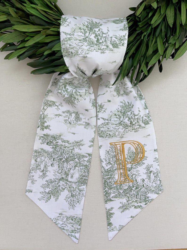 K&ouml;nnte beinhalten: Wei&szlig;er Schal mit gr&uuml;nem Toile-de-Jouy-Muster und einem goldgestickten Buchstaben "P". Der Schal ist &uuml;ber einem gr&uuml;nen Kranz drapiert. Das Toile-Muster zeigt eine l&auml;ndliche Szene.