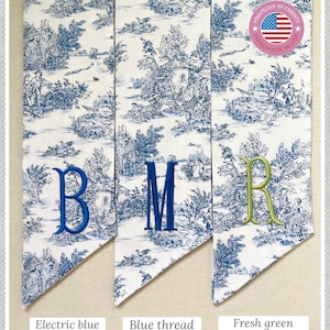 K&ouml;nnte beinhalten: Drei personalisierte Stoffbanner mit gestickten Initialen. Die Banner zeigen ein blau-wei&szlig;es Toile-Muster. Die Initialen sind in elektrisch blauem, blauem und frischem gr&uuml;nem Garn gestickt. Der Text beschreibt die Garnfarbe und den Schriftstil.