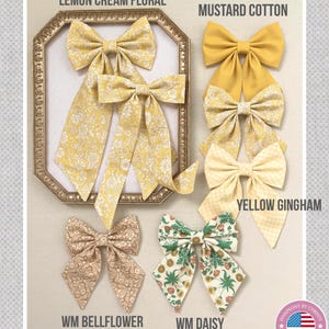 Può includere: Un assortimento di fiocchi in tessuto fatti a mano in vari modelli e colori, tra cui floreale giallo, cotone senape e giallo gingham. I fiocchi sono esposti con il testo "PinkPeony by Christy" e dettagli del prodotto.