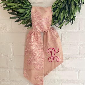 K&ouml;nnte beinhalten: Ein rosa Schal mit Toile-Muster und einem kursiven Initialen-Stick in dunklerem Rosa. Der Schal ist &uuml;ber einem gr&uuml;nen Kranz drapiert, vor einer wei&szlig;en Backsteinmauer.
