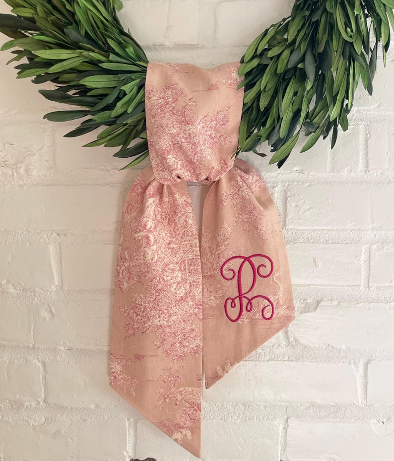 K&ouml;nnte beinhalten: Ein rosa Schal mit Toile-Muster und einem kursiven Initialen-Stick in dunklerem Rosa. Der Schal ist &uuml;ber einem gr&uuml;nen Kranz drapiert, vor einer wei&szlig;en Backsteinmauer.