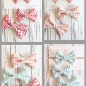 May include: Set of six gingham and striped fabric bow ties in pink, peach, and light blue. The bows are arranged in three pairs on a white wooden surface. The bows are handmade and are perfect for adding a touch of whimsy to any outfit.