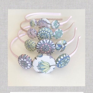 May include: A set of four fabric covered button headbands. The headbands are pink and the buttons are covered in floral fabric with shades of blue, green, pink, and yellow.