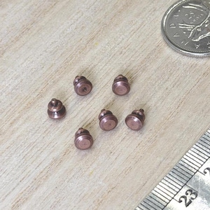 May include: Six small, round, metallic copper-coloured pin backs or fasteners arranged in a cluster. A silver-coloured Canadian coin and a metal ruler showing measurements in centimetres are positioned next to the fasteners for scale reference.