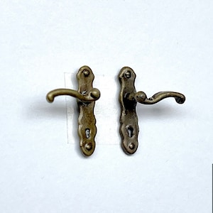 Op de afbeelding: Twee antieke goudkleurige deurgrepen met sierlijke details. Elke greep heeft een gebogen hendel en een sleutelgat. De grepen zijn gemonteerd op een witte achtergrond.
